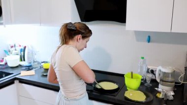 Genç bir kadın şık bir mutfakta krep hazırlıyor. Odaklanmış bir dikkatle, pişirme sürecinin tadını çıkarırken hamuru ustalıkla sıcak tavaya çeviriyor..