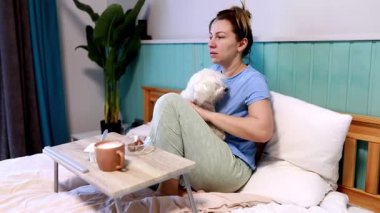 Bir kadın, sıcak bir içecek ve kahvaltı içerken küçük bir köpekle yatakta uykulu bir şekilde oturur. Oda rahatlatıcı bir renk düzeni ve rahat bir atmosfere sahip..