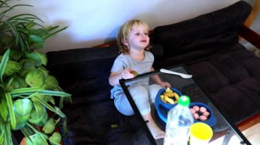 Küçük bir çocuk rahat bir kanepeye oturmuş yapraklı bitkiler ve neşeli bir atmosferle çevrili atıştırmalıkların tadını çıkarırken, parlak kahkahalar odayı dolduruyor..