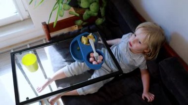 Küçük bir çocuk kanepede dinlenir, yemek dolu mavi bir tepsiden yemek yer. Güneş ışığı pencereden süzülerek odada sıcak bir atmosfer yaratır..