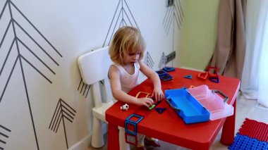 Genç bir çocuk, parlak kırmızı bir masa üzerinde renkli yapı taşları birleştirerek yaratıcı bir oyun oynar. Samimi odada eğlenceli bir duvar dekorasyonu ve hayal gücü var..