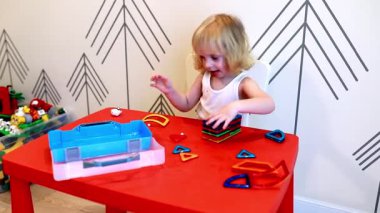 Küçük bir çocuk kırmızı bir masada neşeyle oynar, canlı geometrik şekilleri farklı yapılara ayırır. Oyuncu ortam yaratıcılığı ve hayal gücünü teşvik eder..