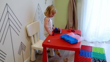 Genç bir çocuk, ferah ve neşeli bir odada renkli oyuncaklarla çevrili parlak kırmızı bir masada oynuyor. Atmosfer yaratıcılığı ve keşfi teşvik eder..