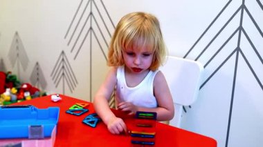 Genç bir çocuk oyuncaklar ve diğer aktivitelerle dolu canlı bir oyun ortamında yapılar inşa ederken yaratıcılık ve odaklanma göstererek renkli manyetik fayanslarla oynar..