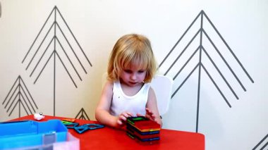 Genç bir çocuk renkli manyetik blokları bir masanın üzerine yığmaya dikkatle odaklanır. Oyuncu ortam parlak renkler ve geometrik şekiller içeriyor, yaratıcılığı ve öğrenmeyi teşvik ediyor.
