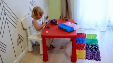 Küçük bir çocuk kırmızı bir masada oturuyor, renkli bloklarla oynuyor ve onları dikkatlice düzenliyor. Oda parlak ve eğlenceli bir dekor içeriyor. Yaratıcılık için davetkar bir atmosfer yaratıyor..