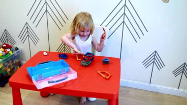 Genç bir çocuk kırmızı bir masada çeşitli renkli yapı taşlarıyla yaratıcı bir oyun oynar. İçerideki canlı ortam, neşeli atmosferi arttırıyor..