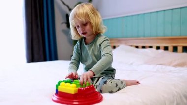 Genç bir çocuk, yumuşak bir yatakta parlak yapı taşlarıyla oynar. Hoş bir yatak odası ortamında şekiller ve yapılar yaratmakla meşguldür..