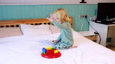 Küçük bir çocuk, sıcak bir odada yatağında otururken kırmızı bir üste renkli yapı taşları birleştirmeye odaklanır. Sabah ışığı boşluğu dolduruyor, neşeli atmosferi güçlendiriyor..