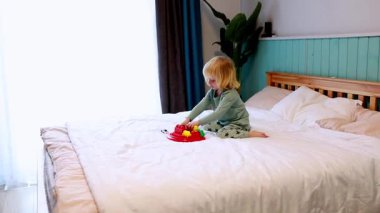 Genç bir çocuk, canlı oyuncaklarla oynaşan bir yatakta oturuyor. Oda, yakınlardaki bir pencereden gelen yumuşak doğal ışık ile sıcak bir atmosfer yaratıyor..