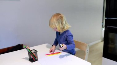 Genç bir çocuk renkli kalemler ve kağıt kullanarak bir masada bir çizim oturumuna katılıyor. Atmosfer parlak ve davetkar, yaratıcılığı ve hayal gücünü teşvik ediyor..