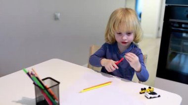 Küçük çocuk, mutfak masasında parlak kalemlerle boyamaya odaklanıyor, gün boyunca rahat bir ev ortamında yaratıcılık ve konsantrasyon gösteriyor..