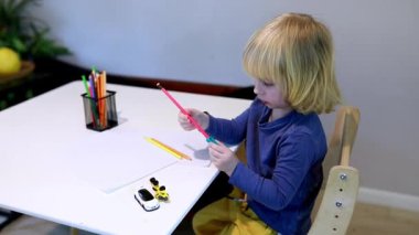 Bir çocuk çizim projesine odaklanmış bir masada oturuyor, çeşitli renkli kalemleri keşfediyor. Çevre parlak ve davetkar, yaratıcılığı ve hayal gücünü destekliyor..