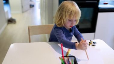 Genç bir çocuk renkli kalemler ve kağıtlarla çevrili olarak dikkatle çizime odaklanır. Etkinlik, yaratıcılık ve konsantrasyonun gösterildiği iyi aydınlatılmış bir mutfakta gerçekleşiyor..