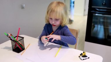 Küçük bir çocuk renkli kalemlerle resim yapmaya odaklanmış bir masada oturuyor. Parlak çalışma alanı, yaratıcılık için eğlenceli bir ortam yaratmak amacıyla etrafa dağılmış kağıt ve sanat malzemeleri barındırıyor..