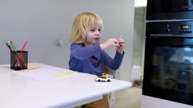 Genç bir çocuk modern bir mutfakta masada oturur, küçük bir oyuncak arabayla oynarken pastel boyalarla resim çizer. Çevre neşeli ve doğal ışıkla dolu..