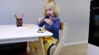 Genç bir çocuk, yaratıcı ifade için renkli kalemler kullanırken oyuncak bir arabayla oynamaya odaklanmış bir masada oturuyor. Kapalı alan parlak ve davetkar..