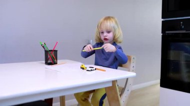 Küçük bir çocuk küçük oyuncak arabalarla oynamaya odaklanır ve güneşli bir mutfakta basit beyaz bir masada kağıt üzerine çizim yapmakla meşgul olur. Renkli kalemler erişilebilir, oyun zamanı artırılıyor.