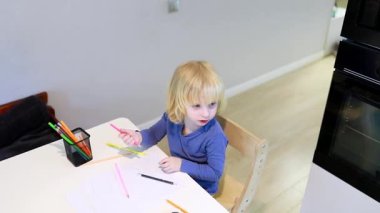 Parlak bir odada, mutfak masasında oturan küçük bir çocuk çeşitli renkli kalemler ve pastel boyalarla çizim yapıyor. Çocuk, yaratıcılık ve hayal gücünü göstermek için sanat yaratmaya odaklanıyor..