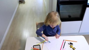 Sarı saçlı küçük bir çocuk renkli kalemler kullanarak dikkatle bir masada resim çizer. Oda parlak ve arka planda bir mutfak alanı görülebilir..