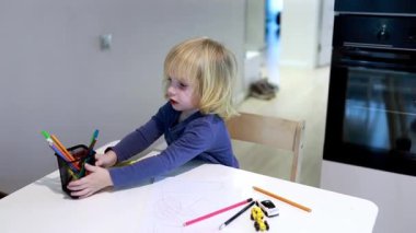 Genç bir çocuk gösterişli bir mutfakta masanın üzerine resim çizerken neşe içinde renkli kalemler düzenler. Bu an, ev ortamında yaratıcılığı ve oyunculuğu gösteriyor..