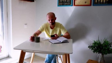Günlük giysiler giymiş bir adam masada bir fincan kahveden hoşlanır, iyi ışıklandırılmış, rahat bir alanda bitkilerle ve dekorla cep telefonuna odaklanır..