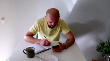 Bir adam sabah rutinini bir masada oturarak yapmayı sever. Yanında bir fincan kahveyle telefonuna odaklanırken bir deftere yazıyor. Güneş ışığı nazikçe mekanı aydınlatır..