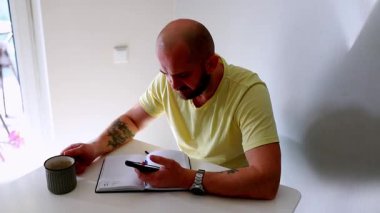 Bir adam telefonuna odaklanırken bir fincan kahveden zevk alır ve parlak bir mutfak masasına notlar yazarak huzurlu bir sabah ortamı yaratır..