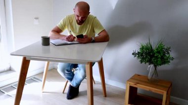 Modern bir masadaki not defterine notlar yazarken bir adam akıllı telefonuyla meşgul oluyor. Güneş ışığı kapalı alandaki minimalist ortamı aydınlatır.