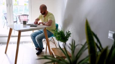 Bir birey, güneşli ve çağdaş bir dairede minimalist bir masada oturuyor. Yeşil bitkiler uzaya doğanın bir dokunuşunu ekler.