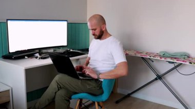 Geniş bir monitör yakınlarda bilgi gösterirken, bir kişi ev ofisinin masasında oturup laptopuna odaklanıyor. Arka planda bir ütü tahtası görünür.
