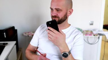 Sakallı bir adam akıllı telefonunu kullanarak canlı bir konuşma yapar. Rahat, aydınlık, modern bir yaşam alanında oturuyor, rahat bir tavır sergiliyor..