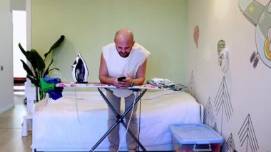 Bir adam akıllı telefonuna bakarken ütü masasında giysileri ütülüyor. Parlak, modern odada bir yatak ve bitkiler var. Rahatlamış bir atmosfer yaratıyor. Çoklu görev için mükemmel..