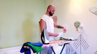 Parlak bir odadaki ütü masasının başında duran bir adam, canlı duvar süslemeleri ve doğal ışık arasında giysileri düzenlemeye ve ütülemeye odaklanıyor..