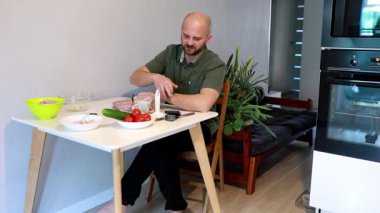 Bir adam mutfak masasında oturur sebze keser ve yemek için malzemeler hazırlar. Modern mutfakta bitkiler ve basit bir dekor var. Sıcak bir atmosfer yaratıyor..