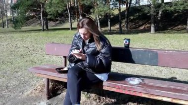Parktaki bankta oturan bir kadın yemeğin tadını çıkarıyor ve akıllı telefonuyla meşgul oluyor. Güneşli gün, dingin çevrenin ve yemyeşil yeşilliğin altını çizer.