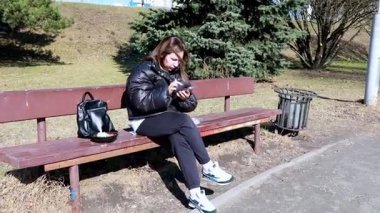 Kabarık ceketli genç bir kadın telefonuna dalmışken bankta oturup havanın tadını çıkarıyor. Park sessiz, etrafta az insan ve parlak güneş var..