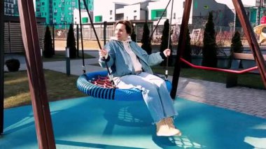 Çağdaş bir çocuk parkında büyük bir salıncakta sallanmaktan hoşlanan bir insan. Çevreleyen yeşillik ve modern mimari hoş bir atmosfer yaratır. Gün aydınlık ve davetkar.