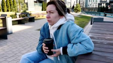 Genç bir kadın modern bir şehir parkında, ahşap bir bankta tek başına oturup fincandan kahve yudumluyor. Parlak bir gökyüzünün altında etrafındakileri düşününce dalgın görünüyor..