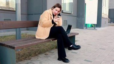 Hafif ceket giymiş bir kadın, çağdaş bir binanın dışındaki bankta oturuyor ve akıllı telefonuna bakarken şaşkınlığını dile getiriyor. Bilgisayarı yakınlarda..