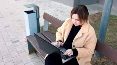 Sonbaharda bir şehir parkında, bir kadın bir bankta otururken dizüstü bilgisayarıyla yazar. Çevrede asfalt yollar ve yeşil çimenler bulunmakta ve sakin bir çalışma ortamı oluşturmaktadır..