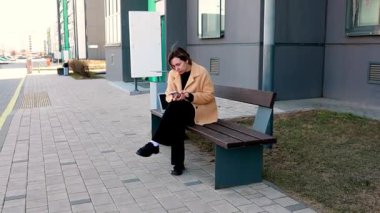 Çağdaş bir binanın önündeki bankta oturan bronz ceketli genç bir kadın akıllı telefonuna dalmıştı. Sahne, berrak gökyüzünün altında sakin bir şehir görüntüsü yakalıyor..