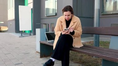 Genç bir kadın, akıllı telefonuna odaklanmış, çağdaş bir binanın dışındaki bankta rahatça oturuyor. Görevine dalmış bir uygulamayla uğraşırken yanında bir dizüstü bilgisayar duruyor..