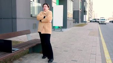 Bir kadın, kürsüde dizüstü bilgisayar kullanırken, sıcak bir palto giymiş, modern bir binanın yanındaki kaldırımda duruyor. Ayarda bir şehir manzarası ve serin bir atmosfer yer alıyor..
