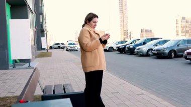 Açık kahverengi paltolu bir kadın kaldırımda duruyor, akıllı telefonuna bakıyor. Modern binalar ve park halindeki arabalar güneşli havada kentsel bir zemin oluşturuyor..