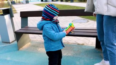 Sıcak giyinmiş genç bir çocuk, parkta oyun oynamaktan zevk alıyor, elinde renkli bir oyuncakla bankın başında dikiliyor. Park ayarları bir oyun alanı gösteriyor.