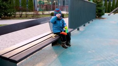 Canlı bir oyun parkındaki bankta oturan küçük bir çocuk sıcak giysiler giyerken oyuncaklarla meşgul oluyor. Çevre canlı, yeşillik ve oyun alanı ekipmanlarıyla donatılmış..