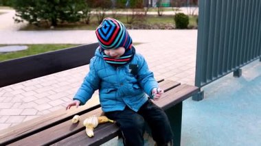 Renkli bir şapkayla sıcacık giyinmiş genç bir çocuk parkta bir bankta mutlu bir şekilde oynuyor, oyuncaklarıyla etkileşime girerken muzdan keyif alıyor, sakin bir manzarayla çevrili..
