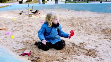 Mavi ceketli küçük bir kız kum havuzunda oturur, oyuncaklarla mutlu bir şekilde oynar ve serin bir öğleden sonra parkta kumu kazar..