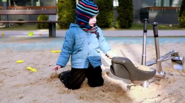 Bir çocuk oyun parkındaki kum havuzunda oturuyor, hevesle kazıyor ve kum oyuncaklarıyla oynuyor. Aydınlık gün ve sıcak hava onu mükemmel bir açık hava aktivitesi yapar..
